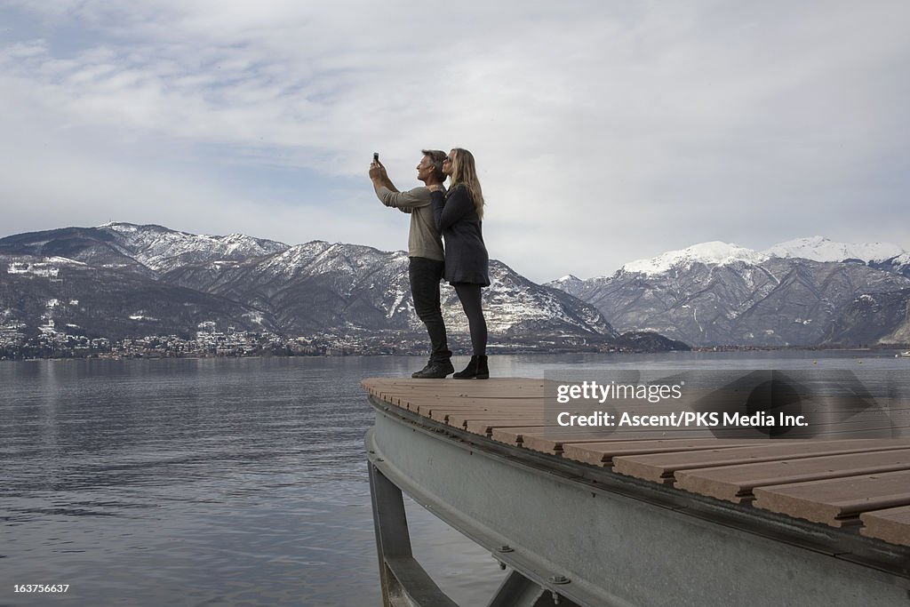 Couple take picture from dock above lake, snowy mt