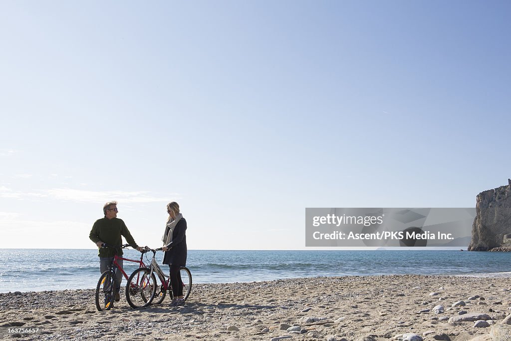 Couple push bikes along sand and pebble beach