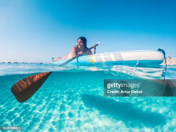 half underwater view of a latin woman lying on the paddleboard rowing in the paradise island of formentera during vacations. - formentera fotografías e imágenes de stock