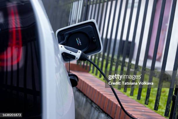 photo of white car with charging equipment plugged in during electric battery charging in backyard - electric car home stock pictures, royalty-free photos & images