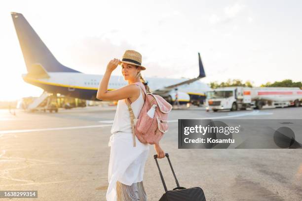 a smiling woman with a backpack on her back is dragging her personal luggage while walking towards the plane - affordable stock pictures, royalty-free photos & images