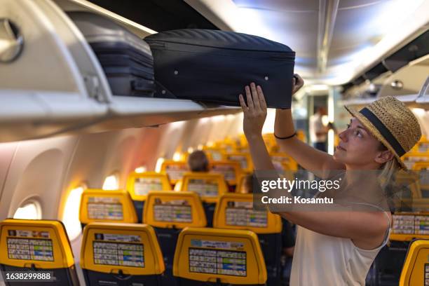 a female tourist puts her suitcase in the overhead compartment of the plane - affordable stock pictures, royalty-free photos & images