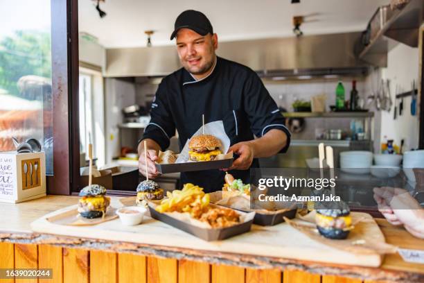 smiling young caucasian chef carring a tray with a delicious big meat burger at a small fast-food restaurant - fastfoodrestaurant stockfoto's en -beelden