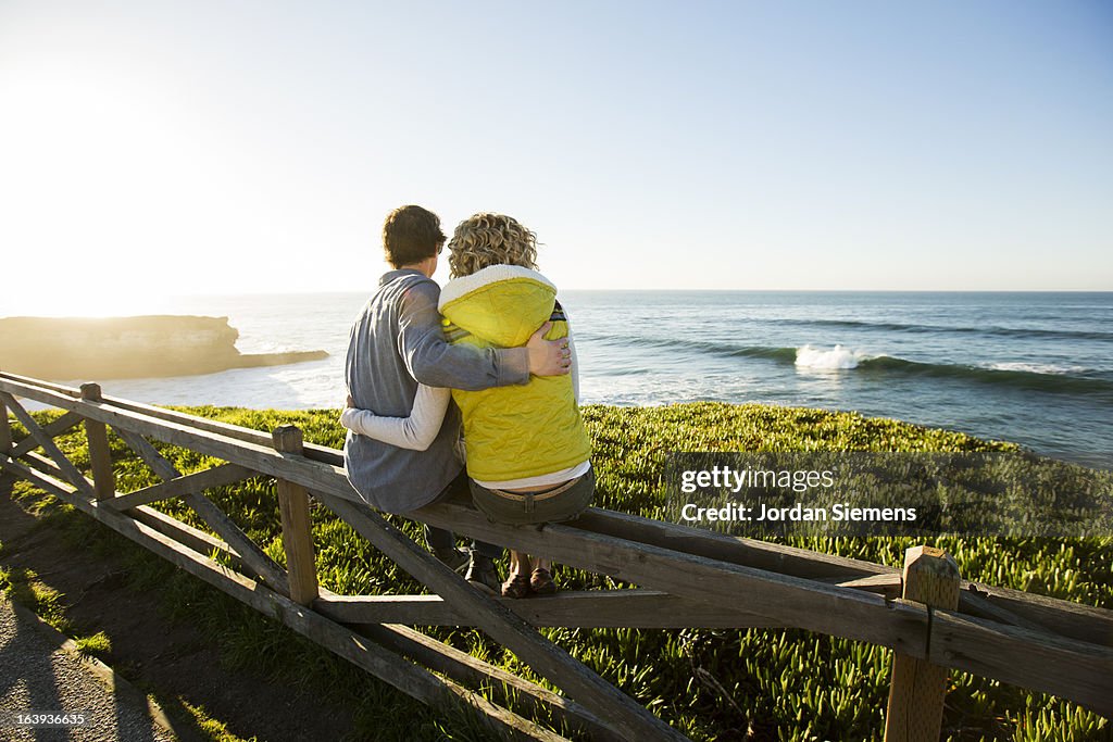 A couple sitting on a fence watching waves.