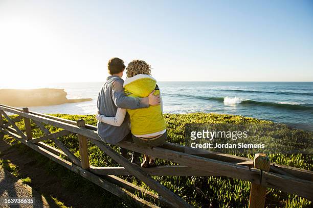 a couple sitting on a fence watching waves. - santa cruz california stock pictures, royalty-free photos & images