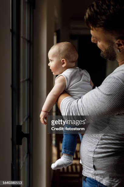 man holding his baby boy, looking out the window together - father baby stock pictures, royalty-free photos & images