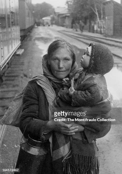 Peasant woman in worn winter clothing holding a young child and metal feed can, standing beside a row of train carriages during World War II; Eastern...