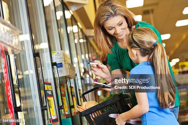 little girl helping mom shop at supermarket with grocery list - inexpensive stockfoto's en -beelden