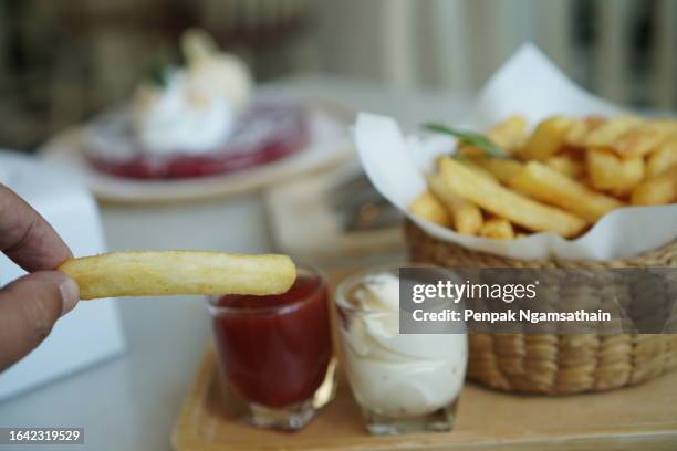 closeup hand eating french fries, potato chips yellow crispy fries in wooden basket with tomato sauce and mayonnaise in clear grass, snack delicious food - condiment stock pictures, royalty-free photos & images