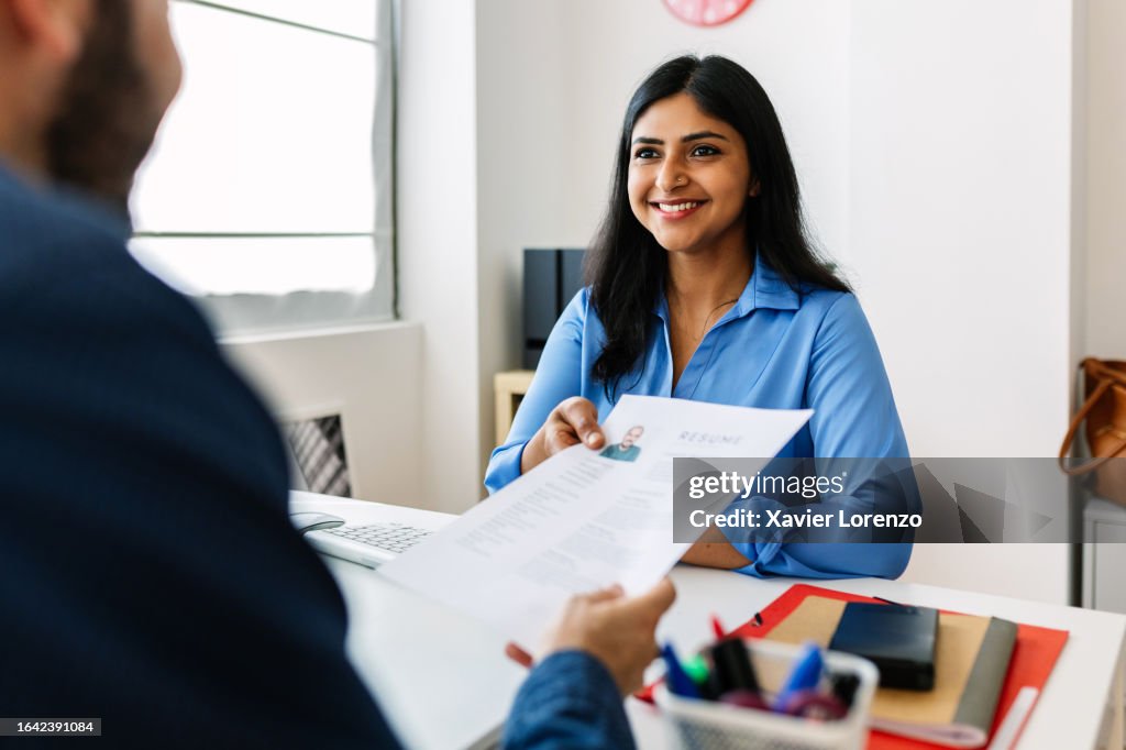 Caucasian job male candidate giving resume to HR manager. Confident successful Indian female recruiter holding resume in hands while having an interview in a modern office, looking and listening to job candidate. Job seeker concept