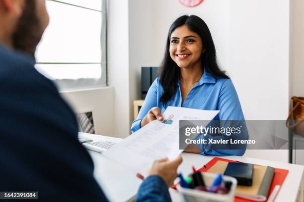 caucasian job male candidate giving resume to hr manager. confident successful indian female recruiter holding resume in hands while having an interview in a modern office, looking and listening to job candidate. job seeker concept - cv bildbanksfoton och bilder