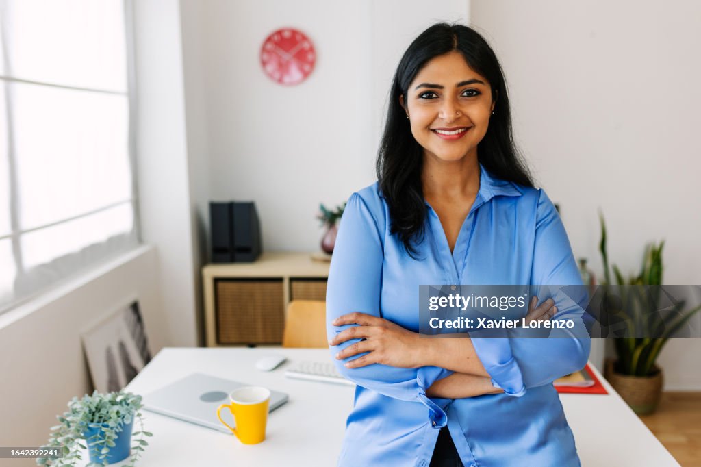 Portrait of smiling Indian businesswoman looking at camera standing at her office. Confident female entrepreneur with crossed arms feeling confident.