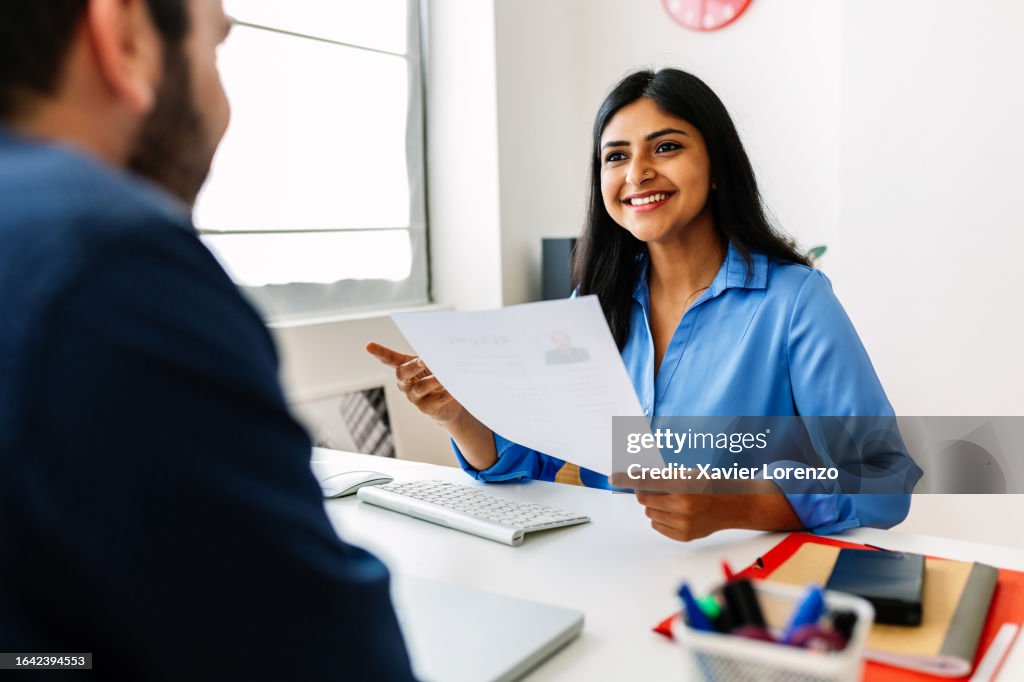 Confident successful Indian female HR recruitment manager holding resume in hands while having an interview in a modern office, looking and listening to job candidate. Friendly interview between HR female director and caucasian job candidate.