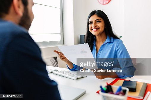 Confident successful Indian female HR recruitment manager holding resume in hands while having an interview in a modern office, looking and listening to job candidate. Friendly interview between HR female director and caucasian job candidate.