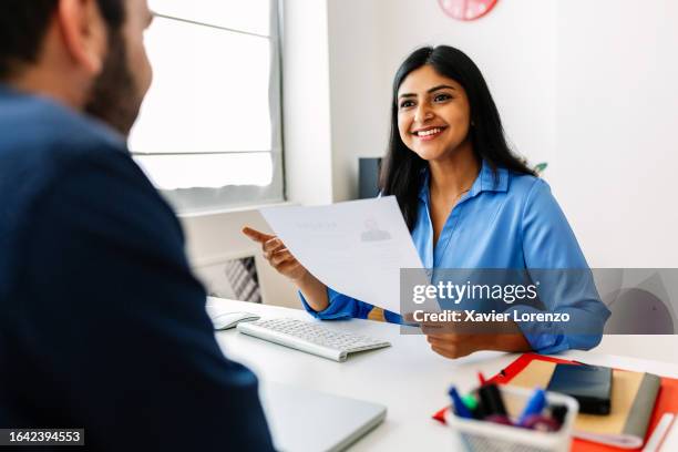 confident successful indian female hr recruitment manager holding resume in hands while having an interview in a modern office, looking and listening to job candidate. friendly interview between hr female director and caucasian job candidate. - recruitment stock pictures, royalty-free photos & images