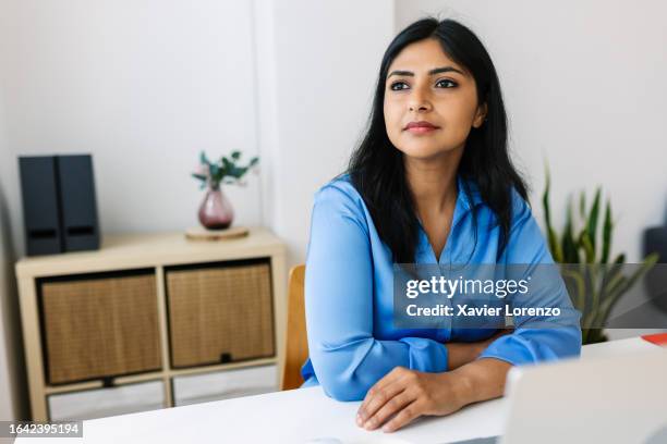 serious mid adult freelance indian businesswoman looking away thinking over creative ideas for project while working at laptop at office. ethnic professional female taking a break sitting at desk looking thoughtfully to the side. - empleada administrativa stock pictures, royalty-free photos & images