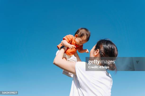 a loving young asian mother lifting her adorable baby girl in the air against blue sky while spending time together in the park. enjoying mother and daughter moment. love and care. family, child and parenthood concept - eltern baby hochheben stock-fotos und bilder