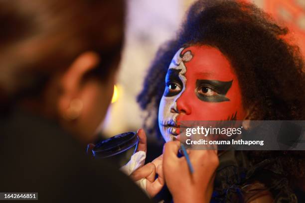 asiatische maskenbildnerin trägt professionelle fettfarbe auf das gesicht des mädchens auf. kriegsbemalung mit blut, narben und wunden im geschmückten wohnzimmer in der halloween-nacht. - halloween zombie makeup stock-fotos und bilder