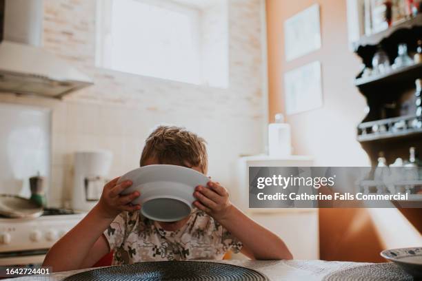 a child drinks milk from a cereal bowl in a domestic kitchen - scodella per zuppa foto e immagini stock