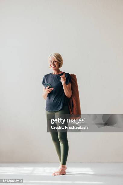 a happy blonde senior woman holding her mobile phone while looking at camera - blonde yoga stock pictures, royalty-free photos & images