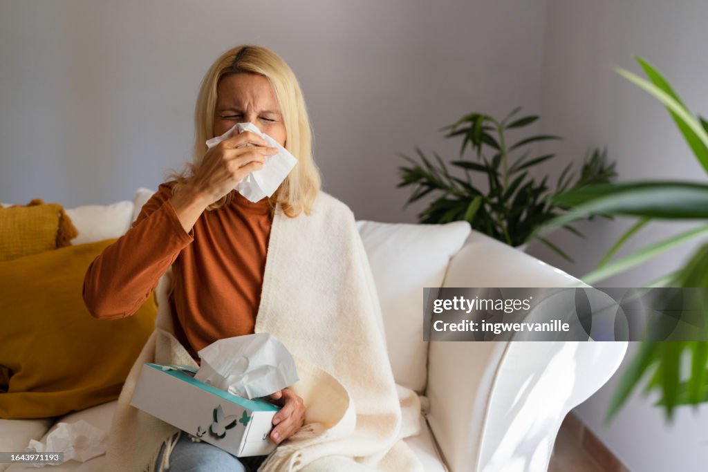 Mature woman sitting on sofa and blowing her nose with a tissue at home