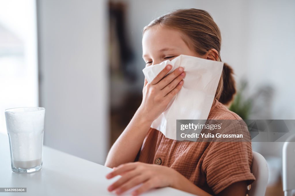 Cute girl funny wipes her face with a paper napkin. In front of her is an empty glass of milk drink.