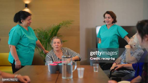 group of senior adult people gathering around table with nurses and enjoying talking with each other in nursing home - kind doctor stock pictures, royalty-free photos & images