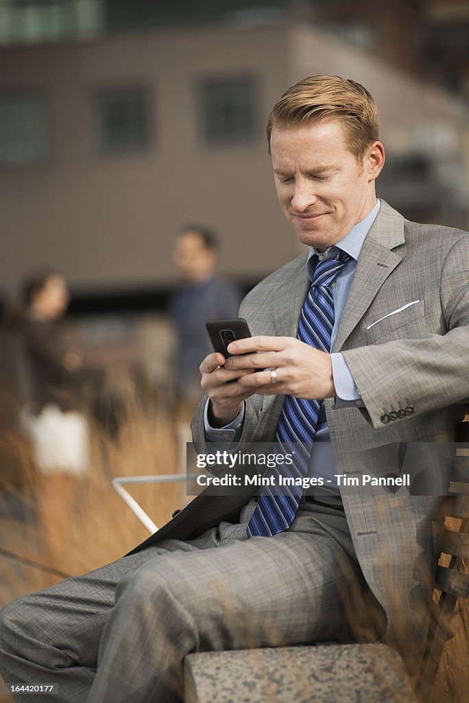 A man sitting on a bench outside a large building,  looking at a cell phone screen or mobile phone.