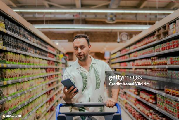 hübscher mann, der nach lebensmitteln aus der liste sucht, ist mobiltelefon - geld verheizen stock-fotos und bilder