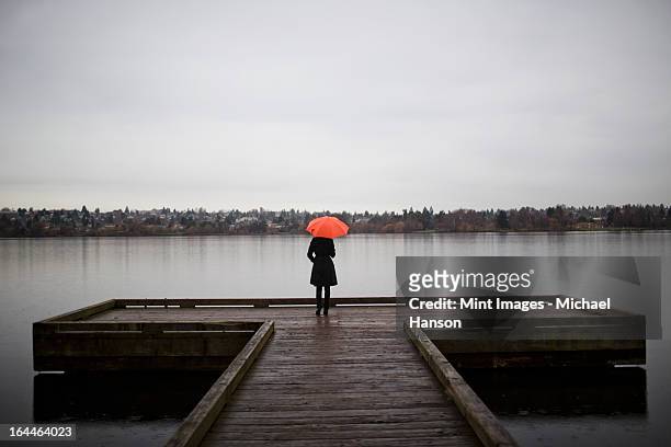 a woman wearing all black and holding an orange umbrella stands on the edge of a dock on a grey and cloudy day in seattle, wa - grey pier stock pictures, royalty-free photos & images