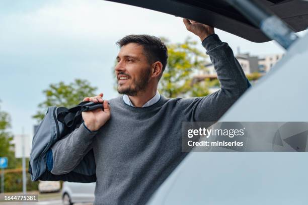 a happy handsome businessman with a sports bag looking away while closing a car trunk - self closing stockfoto's en -beelden