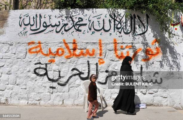 Syrian woman walks with her daughter passed a wall with graffiti written on it, in the northern city of Aleppo on March 24, 2013. Syria's mainstream...