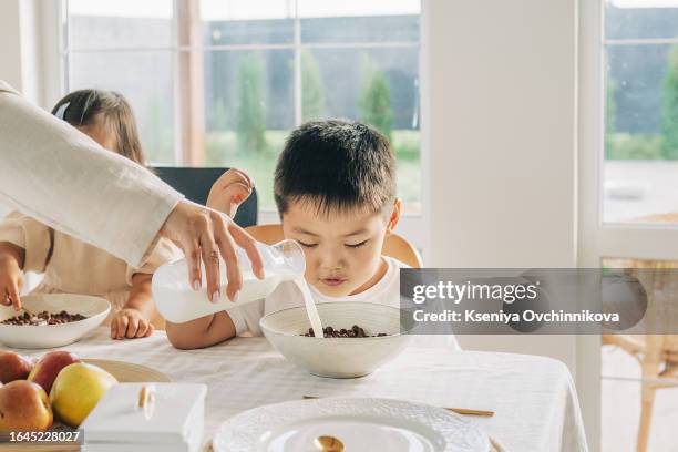 young asian woman pouring calcium drink in glasses and having a bite together with daughter, eating freshly baked biscuits and drinking milk, sitting in kitchen - poured stock pictures, royalty-free photos & images