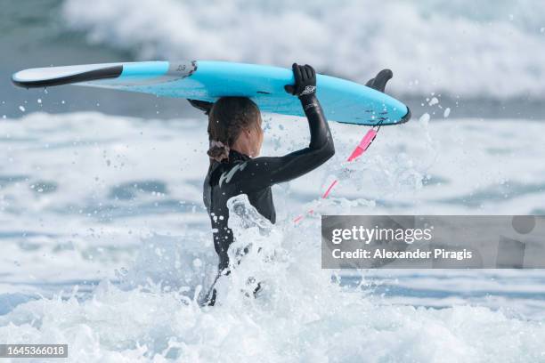 side view of a young female surfer athlete in a wetsuit carrying a surfboard on her head and enters the storm breaking waves of the pacific ocean to go further into the sea - further stock pictures, royalty-free photos & images