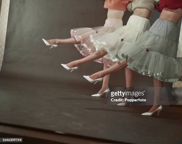Posed studio portrait of three female fashion models wearing multi tiered tulle petticoat underskirts in pastel shades, they stand in a line with...