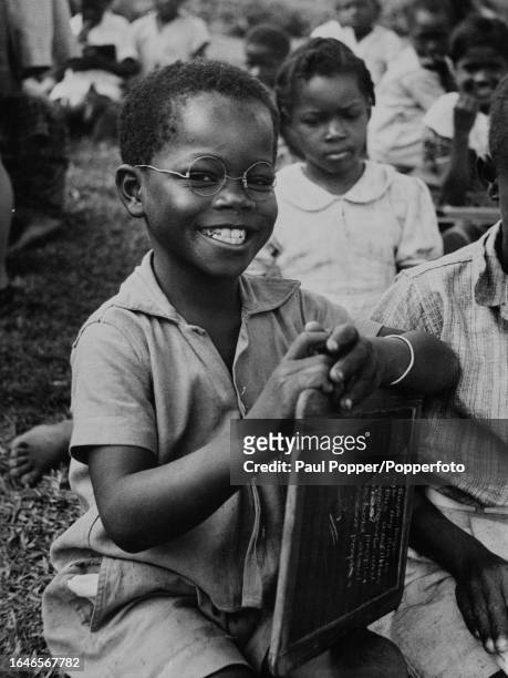 Young boy wearing homemade glasses, without lenses, sits with a small chalkboard held in his lap at a school in Jamaica, circa 1960.