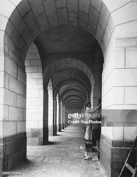 Workman stands on a bench painting a doorway within the archways of the Shell Mex House, also known as 80 Strand, London, circa 1930.