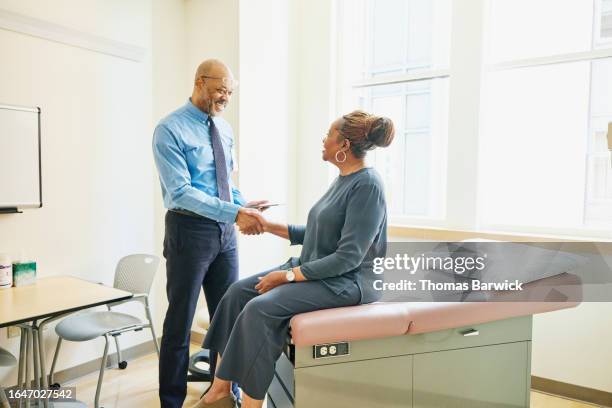 wide shot doctor shaking hands with senior female patient in exam room - atención primaria fotografías e imágenes de stock