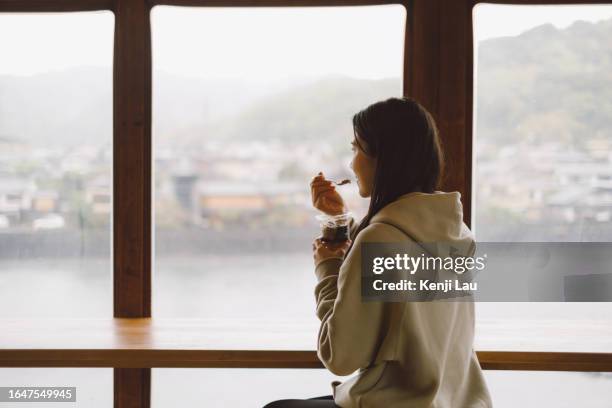 rear view of young asian woman tasting green tea ice cream in japanese cafe and enjoying the secenry of uji throght the window of cafe. concept of foodie, relaxation, travelling, leisure and vacation. - woman looking through ice stock pictures, royalty-free photos & images