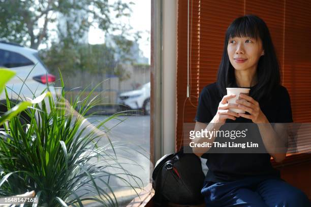 an asian tourist woman holding a cup of coffee by the window in a cafe, she is enjoying the view outdoors - southeast stock pictures, royalty-free photos & images