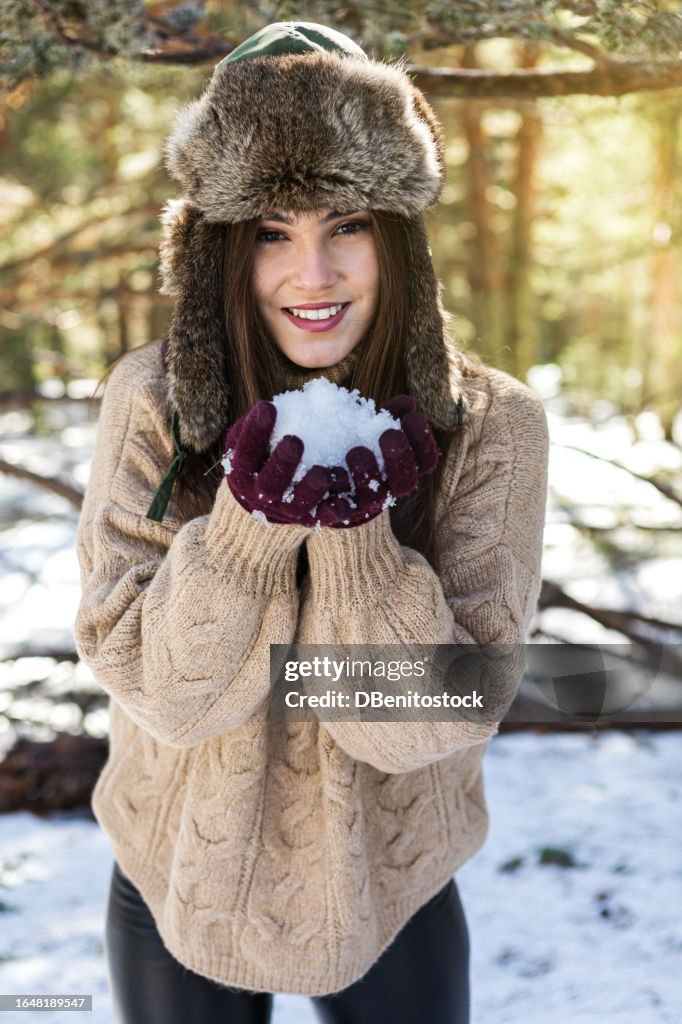 Young black-haired woman, blowing a snowball, in the snowy mountain, wearing a furry hat, gloves and brown wool sweater for winter. Concept of cold, winter, adventure, camping and sky season.