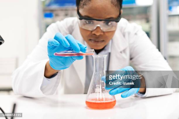 femme scientifique concentrée versant des produits chimiques dans un bécher tout en travaillant en laboratoire - biochimiste photos et images de collection