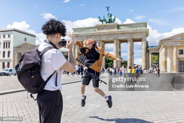 tourist taking pictures of his friend jumping in front of brandenburg gate in berlin - voyageur homme devant monument photos et images de collection