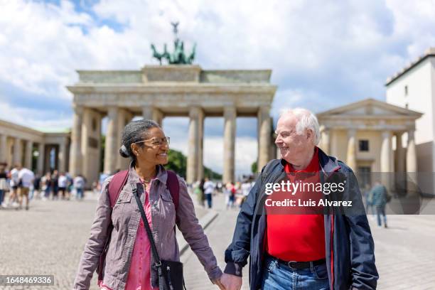 senior couple holding hands walking in front of famous brandenburg gate in berlin - porta de brandemburgo imagens e fotografias de stock