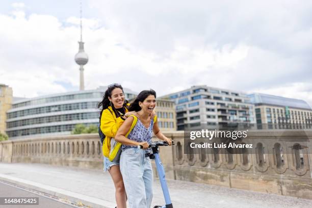 two beautiful young female friends riding on one push scooter in the city - mitte stock pictures, royalty-free photos & images