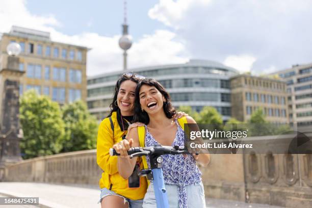 portrait of two cheerful young women friends on holiday having fun on push scooter in city - berlin diversity alexanderplatz stockfoto's en -beelden