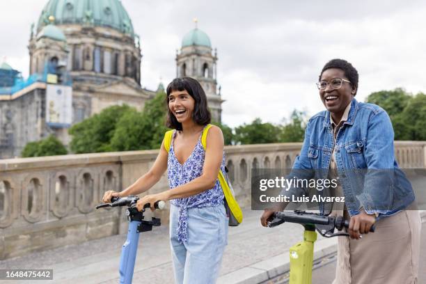 two multiracial women friends on holiday having fun driving electric push scooter in city - roller fahren stock-fotos und bilder