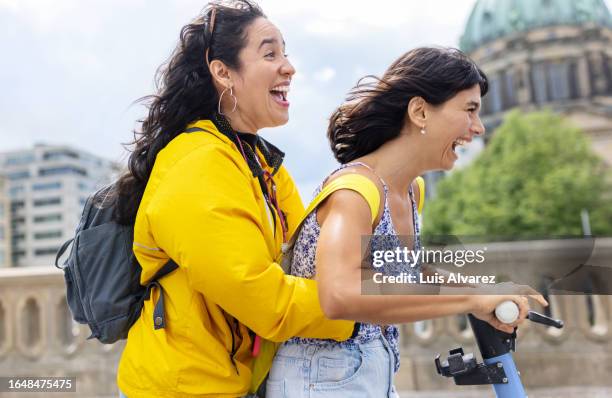 side view of two female friends messing around in a city on electric scooter - mitte stock pictures, royalty-free photos & images