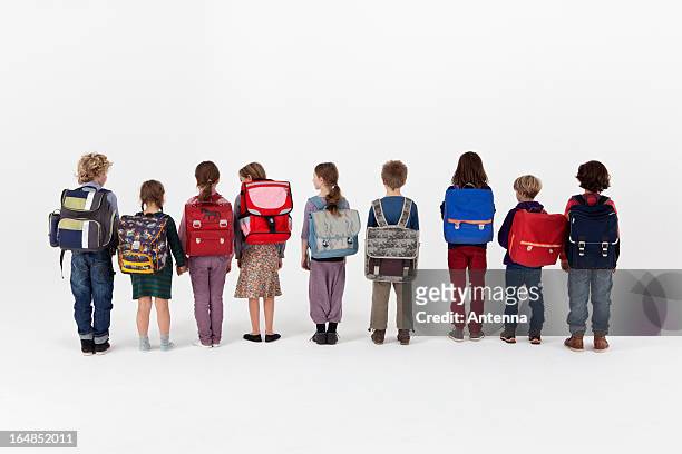 a group of school children wearing backpacks and standing in a row, rear view - schulrucksack stock-fotos und bilder