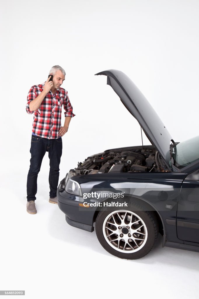 A man standing next to a broke down car while using a cell phone